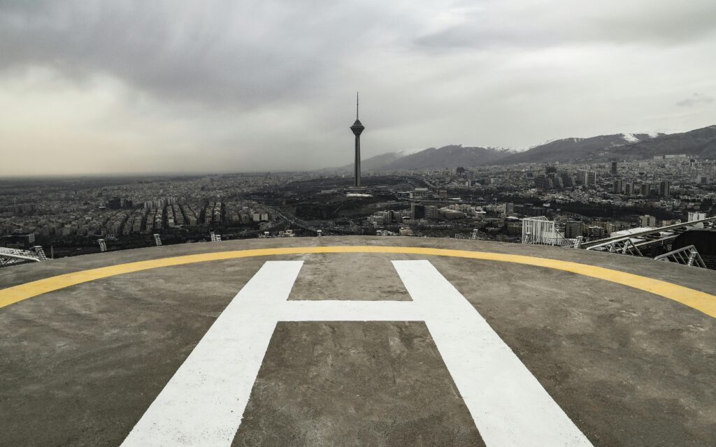 A dramatic view of Tehran's skyline featuring Milad Tower and a helipad under cloudy skies.