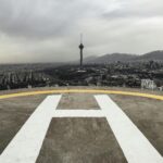 A dramatic view of Tehran's skyline featuring Milad Tower and a helipad under cloudy skies.